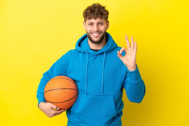 Young handsome caucasian man isolated on yellow background\
playing basketball and making ok sign