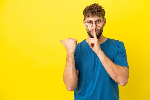 Young handsome caucasian man isolated on yellow background pointing to the side and doing silence gesture