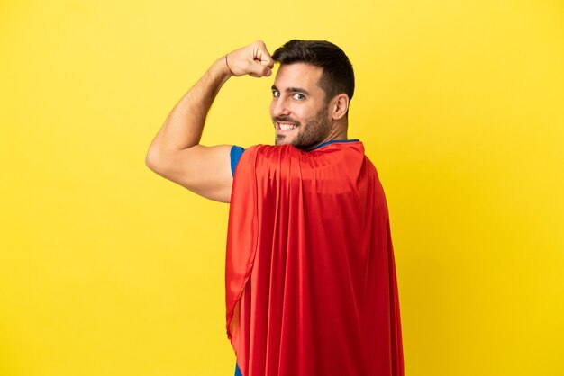 Young handsome caucasian man isolated on yellow background in superhero costume and doing strong gesture