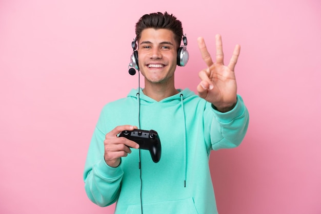 Photo young handsome caucasian man playing with a video game controller isolated on pink background happy and counting three with fingers