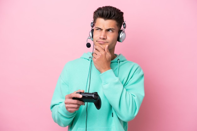 Молодой красивый кавказец играет с контроллером видеоигры на розовом фоне, сомневается и думает