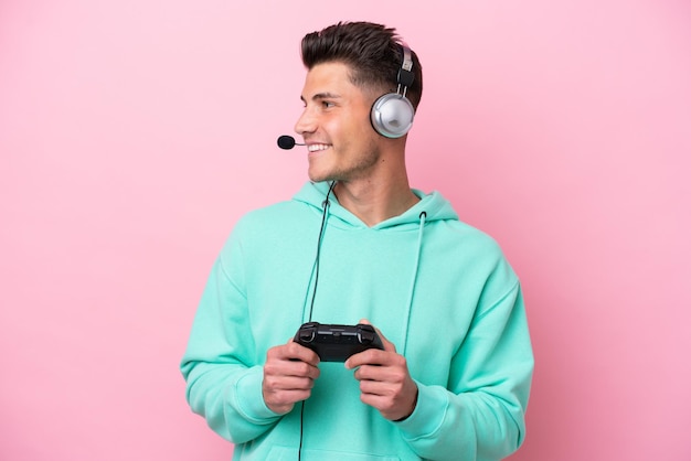 Young handsome caucasian man playing with a video game controller isolated on pink background looking side