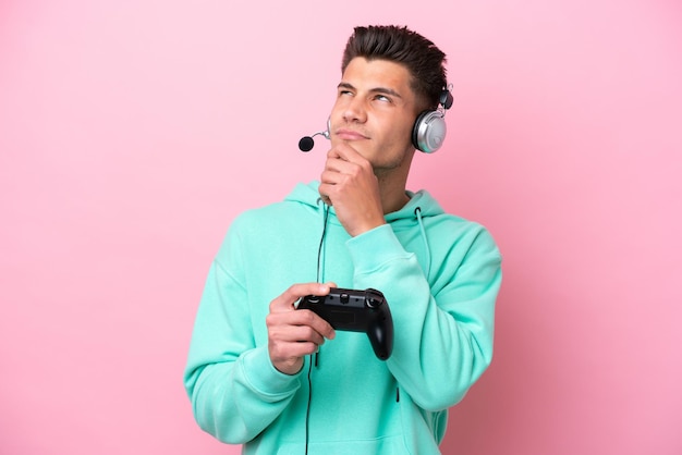 Young handsome caucasian man playing with a video game controller isolated on pink background and looking up