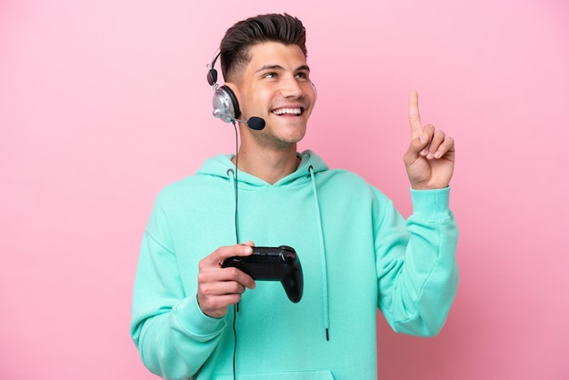 素晴らしいアイデアを指しているピンクの背景に分離されたビデオ ゲーム コントローラーで遊ぶ若いハンサムな白人男性