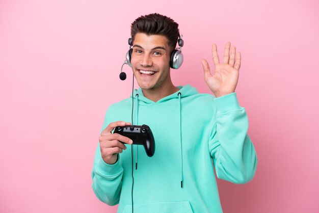 Photo young handsome caucasian man playing with a video game controller isolated on pink background saluting with hand with happy expression