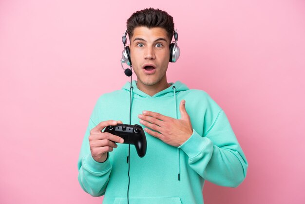 Молодой красивый кавказец, играющий с контроллером видеоигры, изолированным на розовом фоне, удивлен и шокирован, глядя вправо