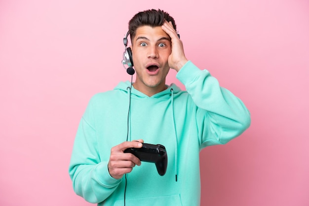 Молодой красивый кавказец играет с контроллером видеоигры на розовом фоне с удивленным выражением лица