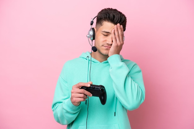 Young handsome caucasian man playing with a video game controller isolated on pink background with tired and sick expression