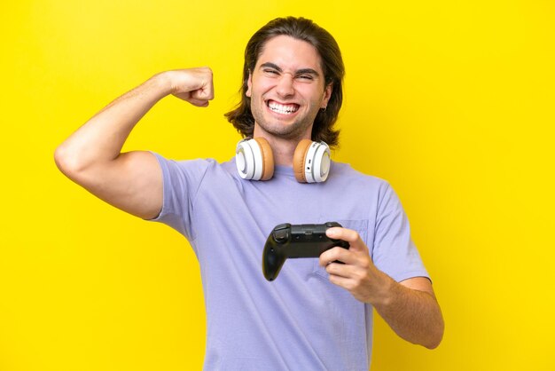 Young handsome caucasian man playing with a video game controller over isolated on yellow background doing strong gesture