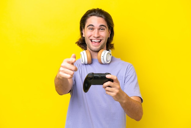 Young handsome caucasian man playing with a video game controller over isolated on yellow background surprised and pointing front