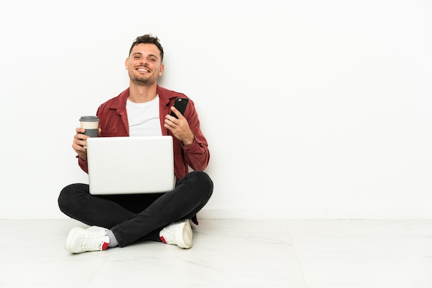 Sit-in di giovane uomo caucasico bello sul pavimento con il computer portatile che tiene il caffè da portare via e un cellulare