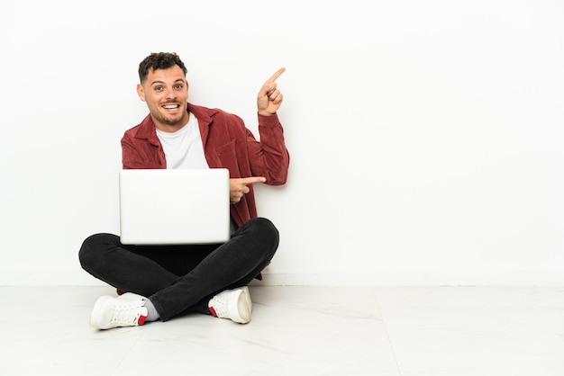 Young handsome caucasian man sit-in on the floor with laptop pointing finger