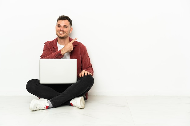 Photo young handsome caucasian man sit-in on the floor with laptop pointing to the side to present a product