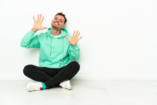 Young handsome caucasian man sitting on the floor counting ten with fingers