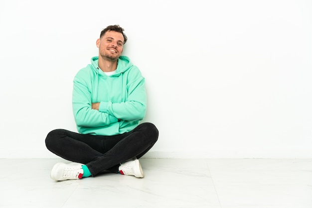 Young handsome caucasian man sitting on the floor with arms crossed and looking forward