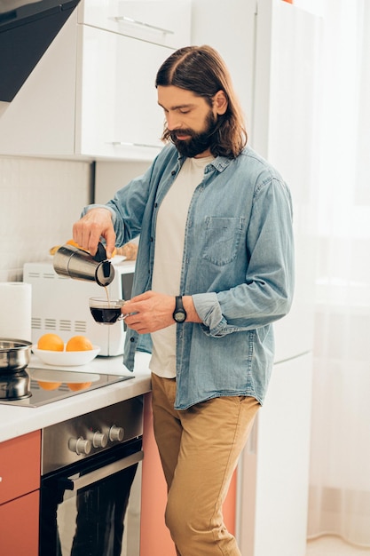 Young handsome Caucasian man standing in the kitchen in casual clothes and pouring coffee