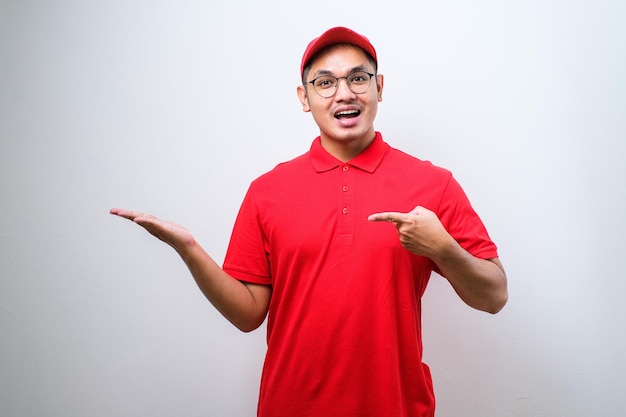 Young handsome chinese delivery man wearing cap standing over isolated white background with a big smile on face pointing with hand and finger to the side looking at the camera