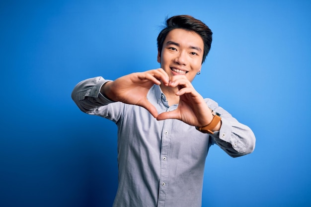 캐주얼 셔츠를 입은 젊고 잘생긴 중국 남자는 격리된 파란색 배경 위에 서서 손으로 심장 기호 모양을 하고 사랑에 미소를 짓고 있습니다.