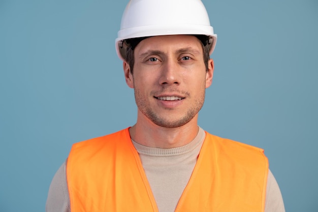 Young handsome civil engineer looking at the camera while standing in studio. Worker wearing hard hat on isolated blue background
