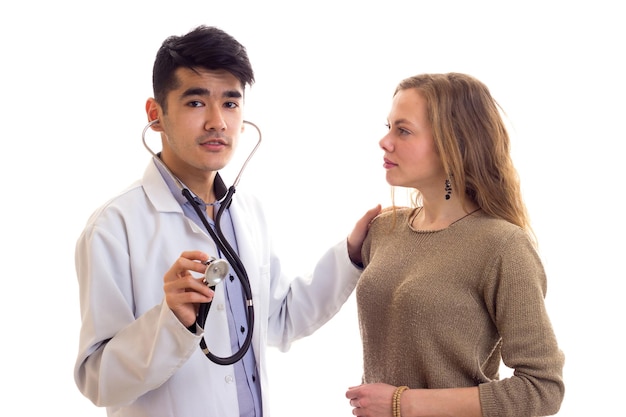 Young handsome doctor in gown with stethoscope listening to the heartbeat of young pretty woman