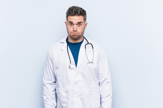 Young handsome doctor man blows cheeks, has tired expression. Facial expression concept.