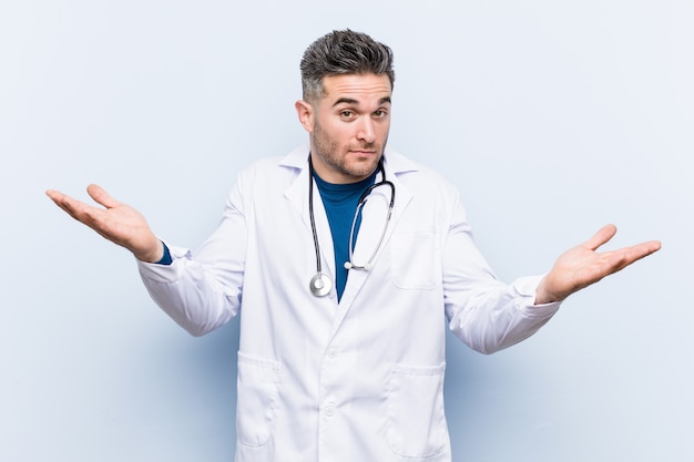 Young handsome doctor man doubting and shrugging shoulders in questioning gesture.