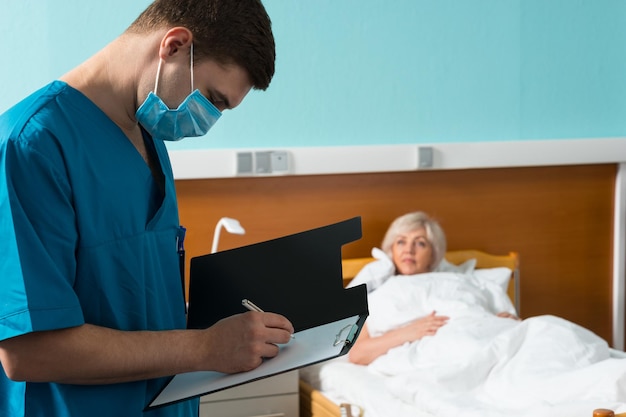 Young handsome doctor in uniform wearing a mask making some notes in report at clipboard while visiting ill patient in the ward. Healthcare concept