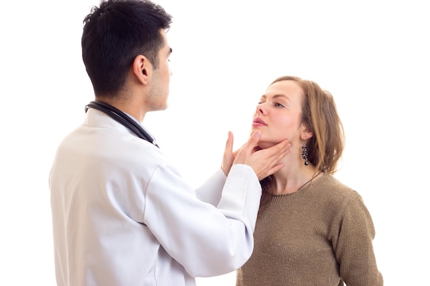 Young handsome doctor with dark hair in white gown with stethoscope on his neck examing young woman