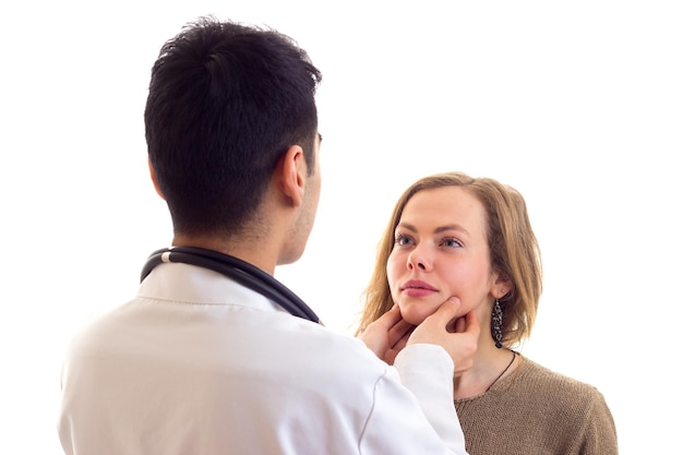 Young handsome doctor with dark hair in white gown with stethoscope on his neck examing young woman