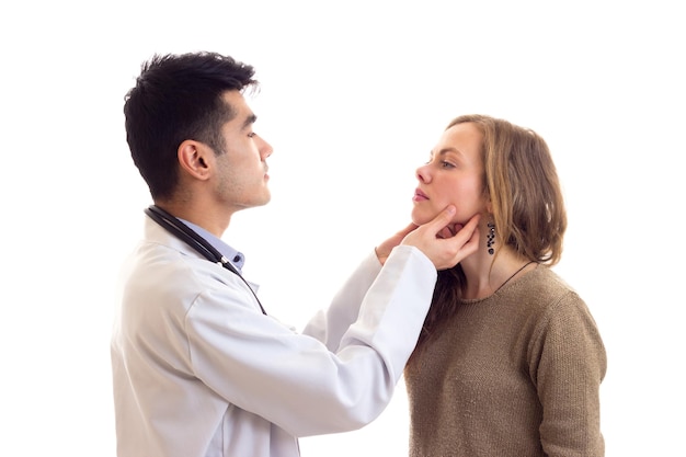 Young handsome doctor with dark hair in white gown with stethoscope on his neck examing young woman