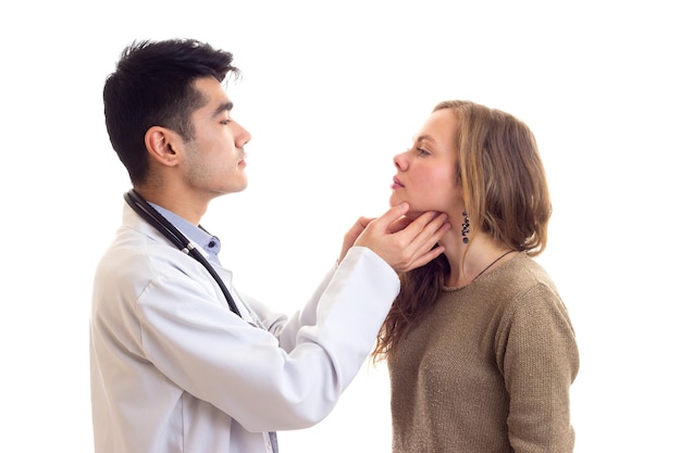 Young handsome doctor with dark hair in white gown with stethoscope on his neck examing young woman