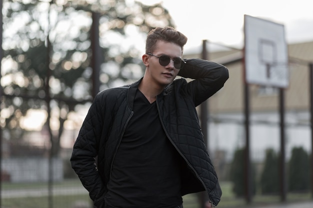 Young handsome fashion man with sunglasses in a black stylish jacket poses at the stadium outdoors