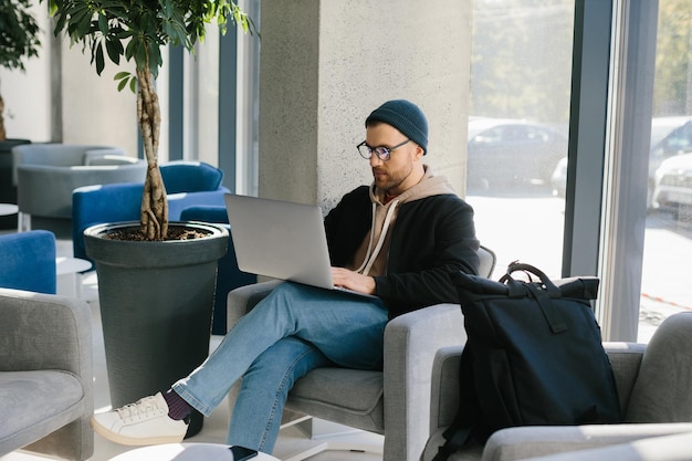 Young handsome freelancer guy working with laptop in office space A man in glasses and a hat is holding a laptop while sitting in a chair in the hall