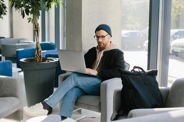 Young handsome freelancer guy working with laptop in office space A man in glasses and a hat is holding a laptop while sitting in a chair in the hall