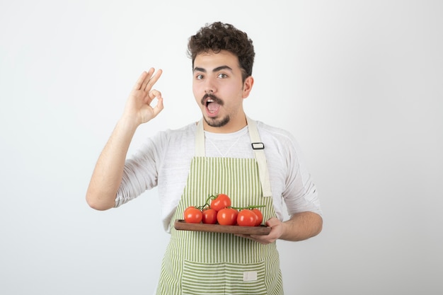 Giovane ragazzo bello che tiene mucchio di pomodori biologici e gesto ok.