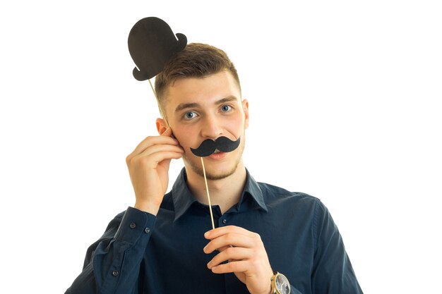 Young handsome guy keeps near the face, paper mustache and a hat isolated on white background