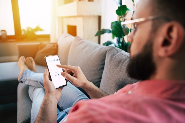 Young handsome happy bearded man in glasses is using smartphone for surfing in net