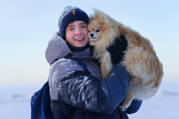 雪の降る冬にペットのポメラニアン スピッツ犬と一緒に歩く手をつないでいる若いハンサムな幸せな男