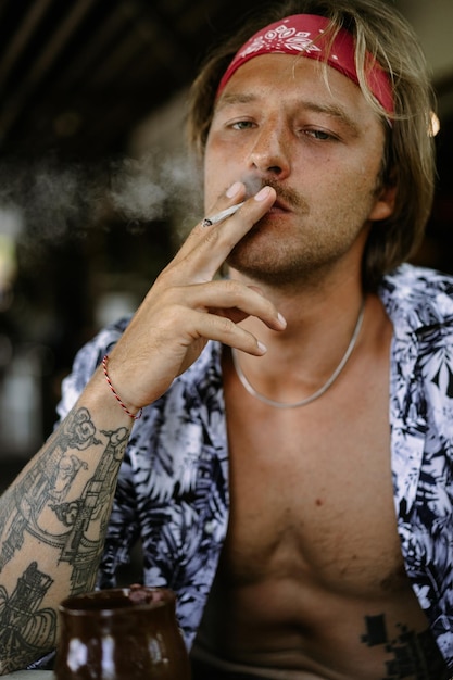 A young handsome hippie man in a red bandana with a bare torso rolls a tobacco cigarette in a cafe in Bali.