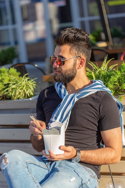 Ragazzo giovane hipster bello mangiare spaghetti cinesi da un pranzo al sacco mentre era seduto su una panchina del parco