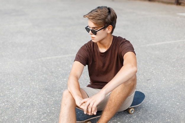 Young handsome hipster male in trendy sunglasses in stylish t-shirt is resting on a skateboard on the basketball court on a summer day