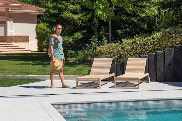 Фото Молодой красивый хипстер, стоящий перед бассейном летом и держащий соломенную шляпу от солнца