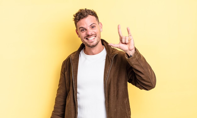 Young handsome hispanic man feeling happy, fun, confident,
positive and rebellious, making rock or heavy metal sign with
hand