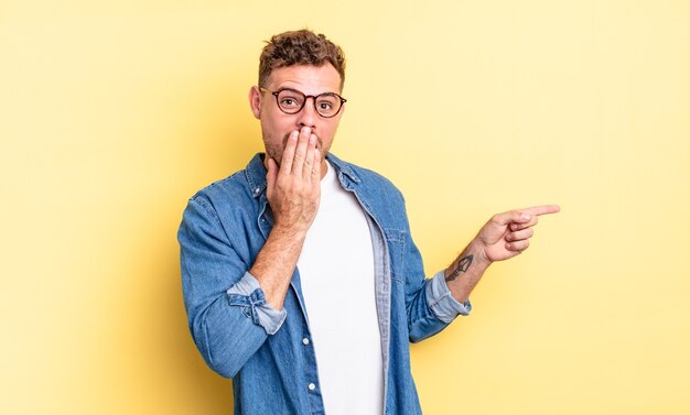 Young handsome hispanic man feeling happy, shocked and surprised, covering mouth with hand and pointing to lateral copy space