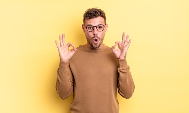 Young handsome hispanic man feeling shocked, amazed and surprised, showing approval making okay sign with both hands