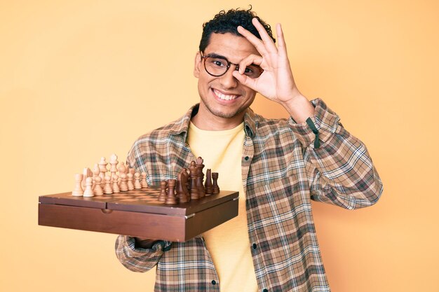 Молодой красивый латиноамериканец, держащий шахматную доску в очках, счастливо улыбающийся, делая знак "ок" с рукой на глазу, глядя сквозь пальцы