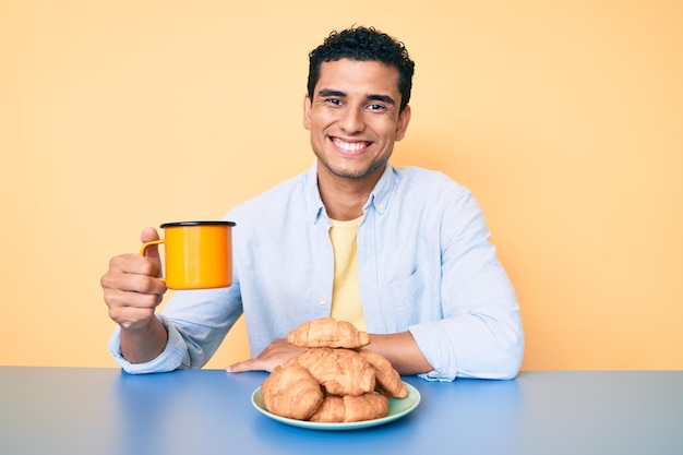 젊고 잘생긴 히스패닉 남자가 긍정적이고 행복한 표정으로 아침 식사를 하고 테이블에 앉아 치아를 보여주는 자신감 있는 미소로 웃고 있습니다.