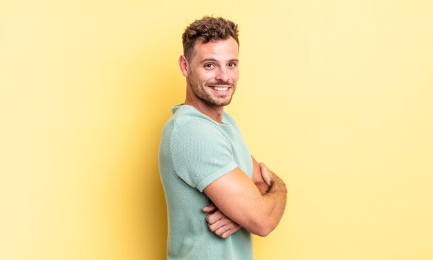 young handsome hispanic man smiling gleefully, feeling happy, satisfied and relaxed, with crossed arms and looking to the side