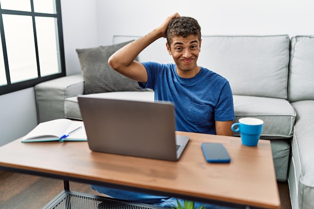 Young handsome hispanic man using laptop sitting on the floor confuse and wonder about question. uncertain with doubt, thinking with hand on head. pensive concept.