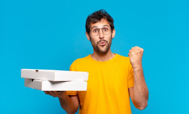 Young handsome indian man celebrating successful a victory and holding take away pizzas